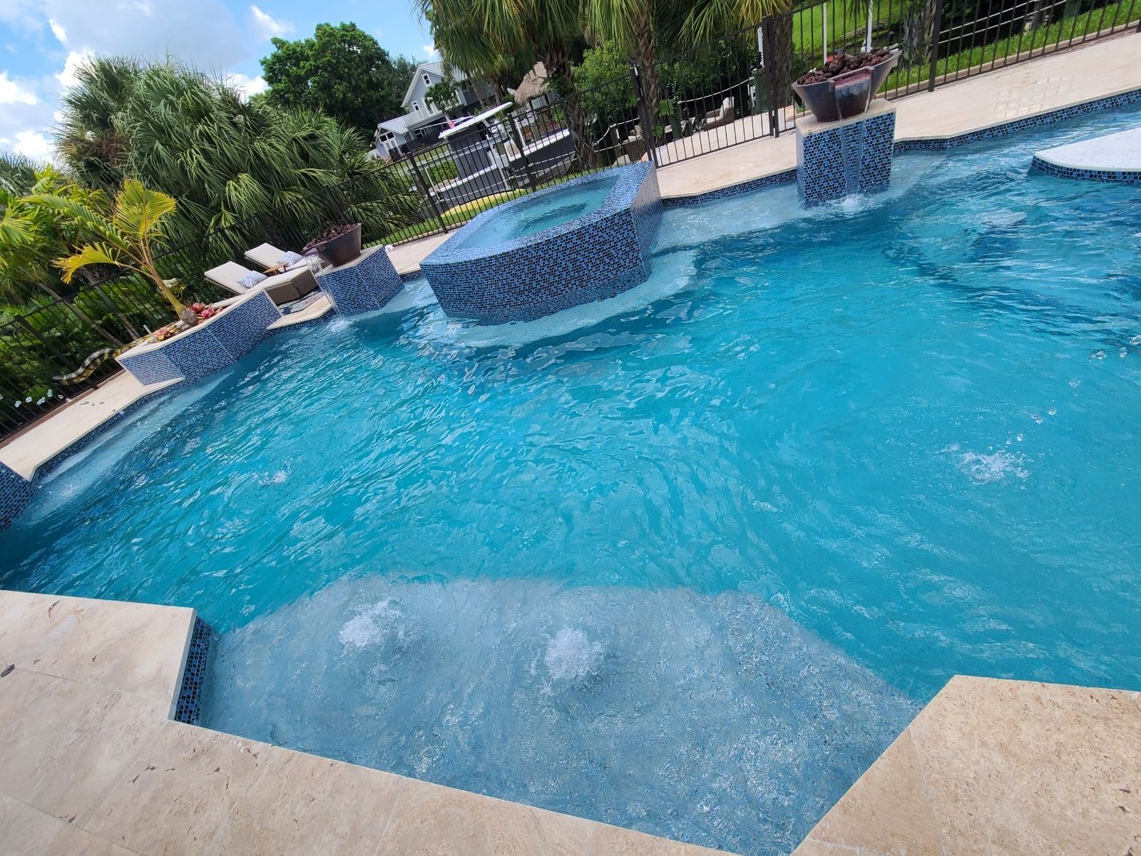Swimming pool with blue water, jacuzzi, and waterfalls. Concrete steps and sunny outdoor setting.