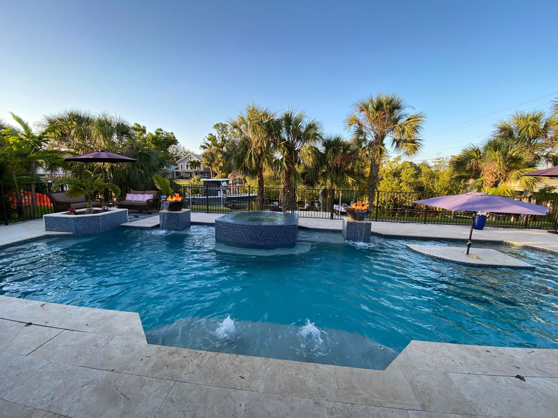 Swimming pool with blue water, surrounded by a stone deck. Palm trees and umbrellas offer shade.