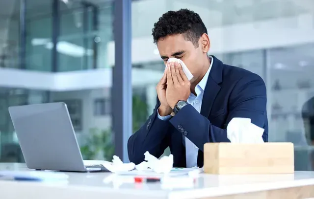 A man is blowing his nose while sitting at a desk with a laptop.