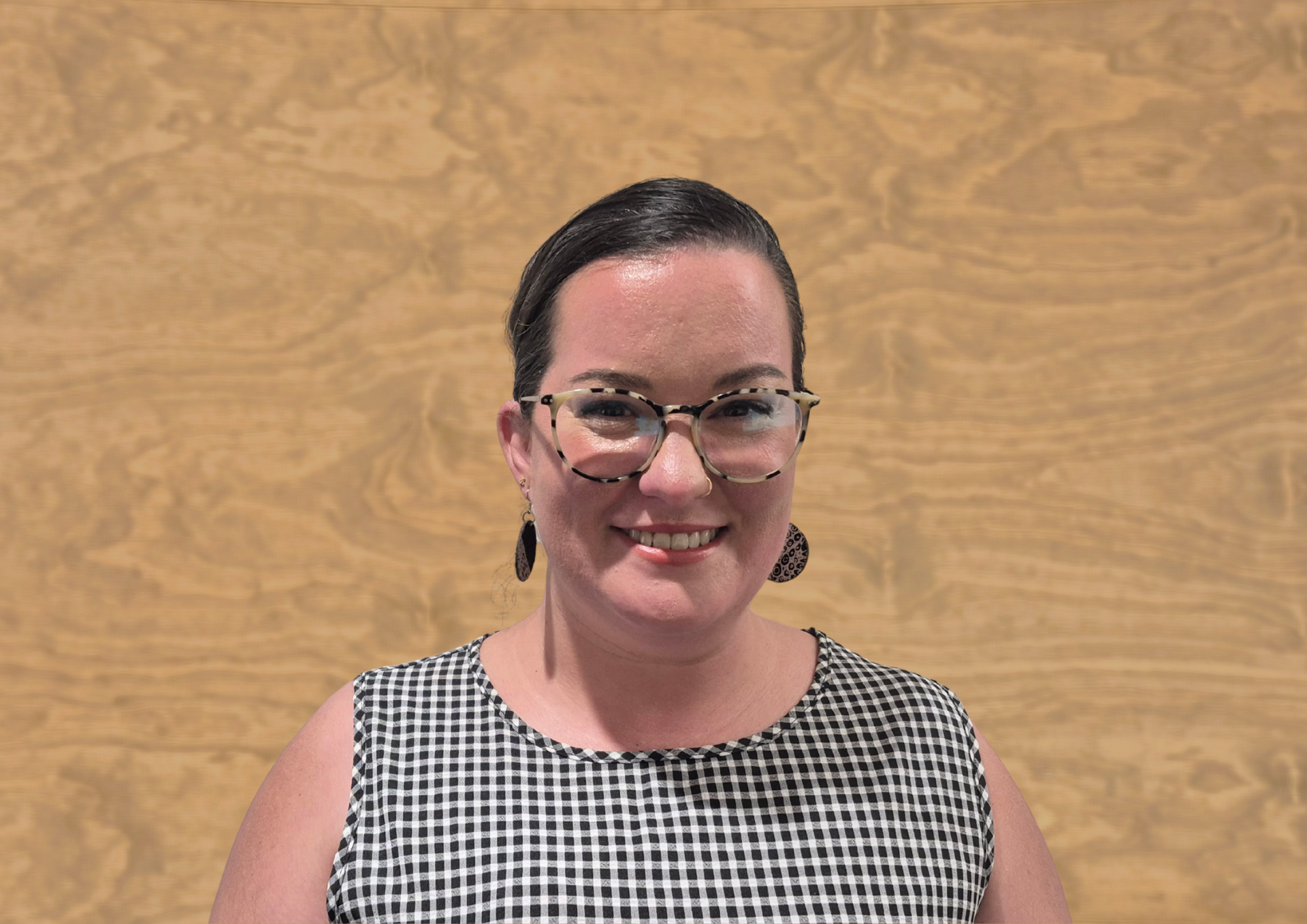 A person with dark hair wearing glasses and a checkered top smiling against a light-brown wooden background.