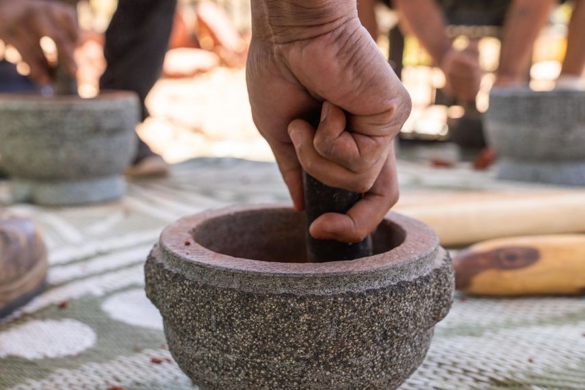 A close-up of a person using a stone pestle to grind ingredients in a stone mortar, set on a textured mat.