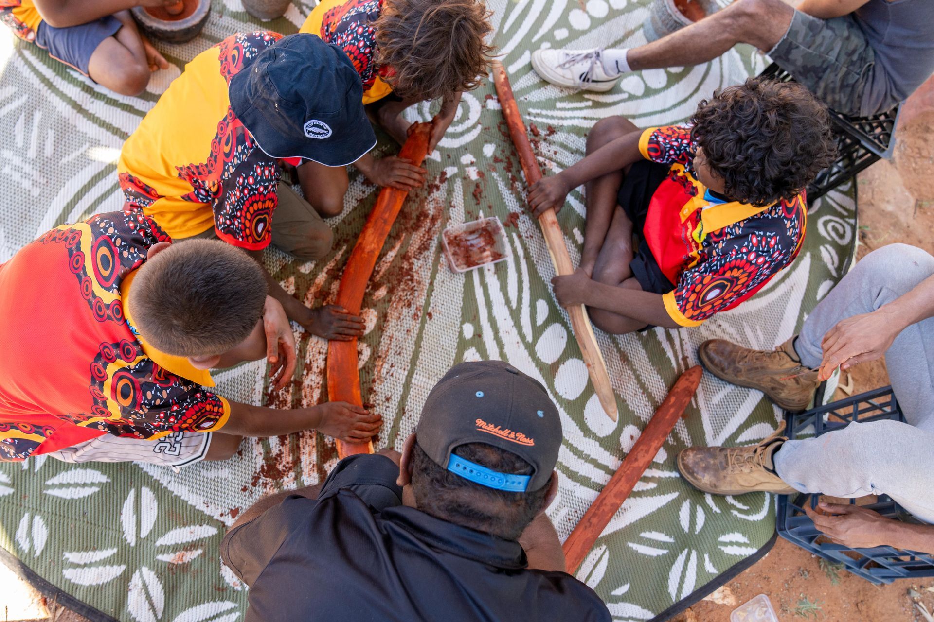 People sit around a patterned mat, working with long wooden tools on a substance, dressed in vibrant patterned shirts.