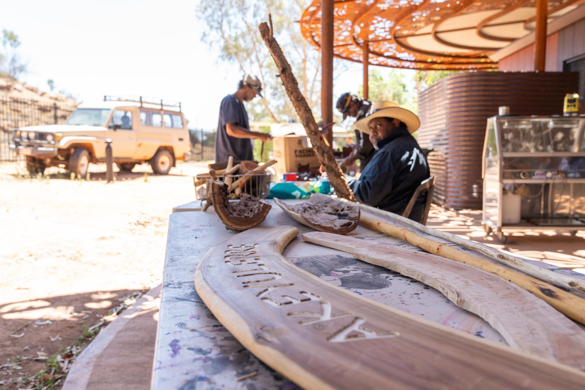 Two people work on wood carving projects at a table outdoors, with a parked vehicle visible in the background.