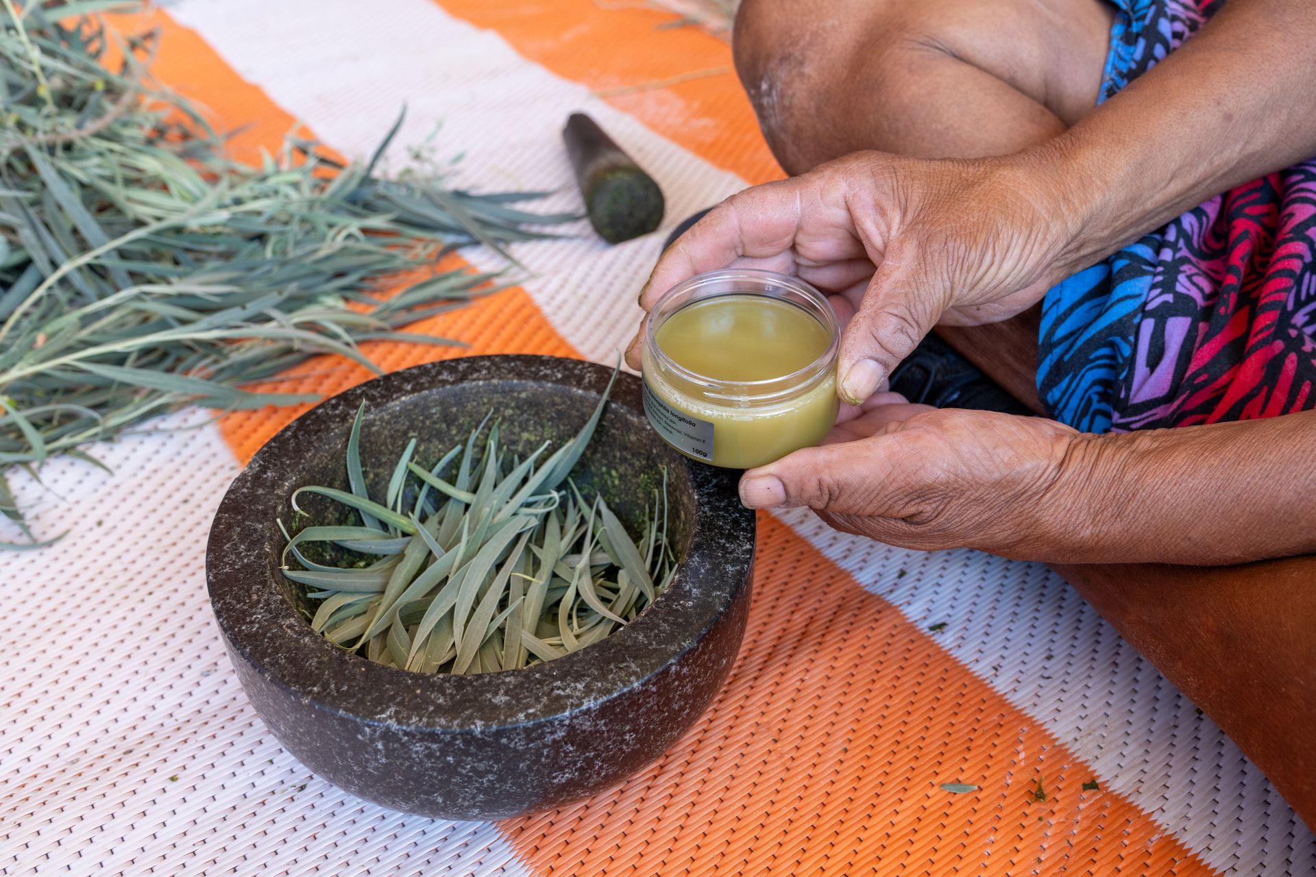 A person’s hands hold a small jar of herbal salve beside a mortar filled with green leaves on an orange-striped cloth.