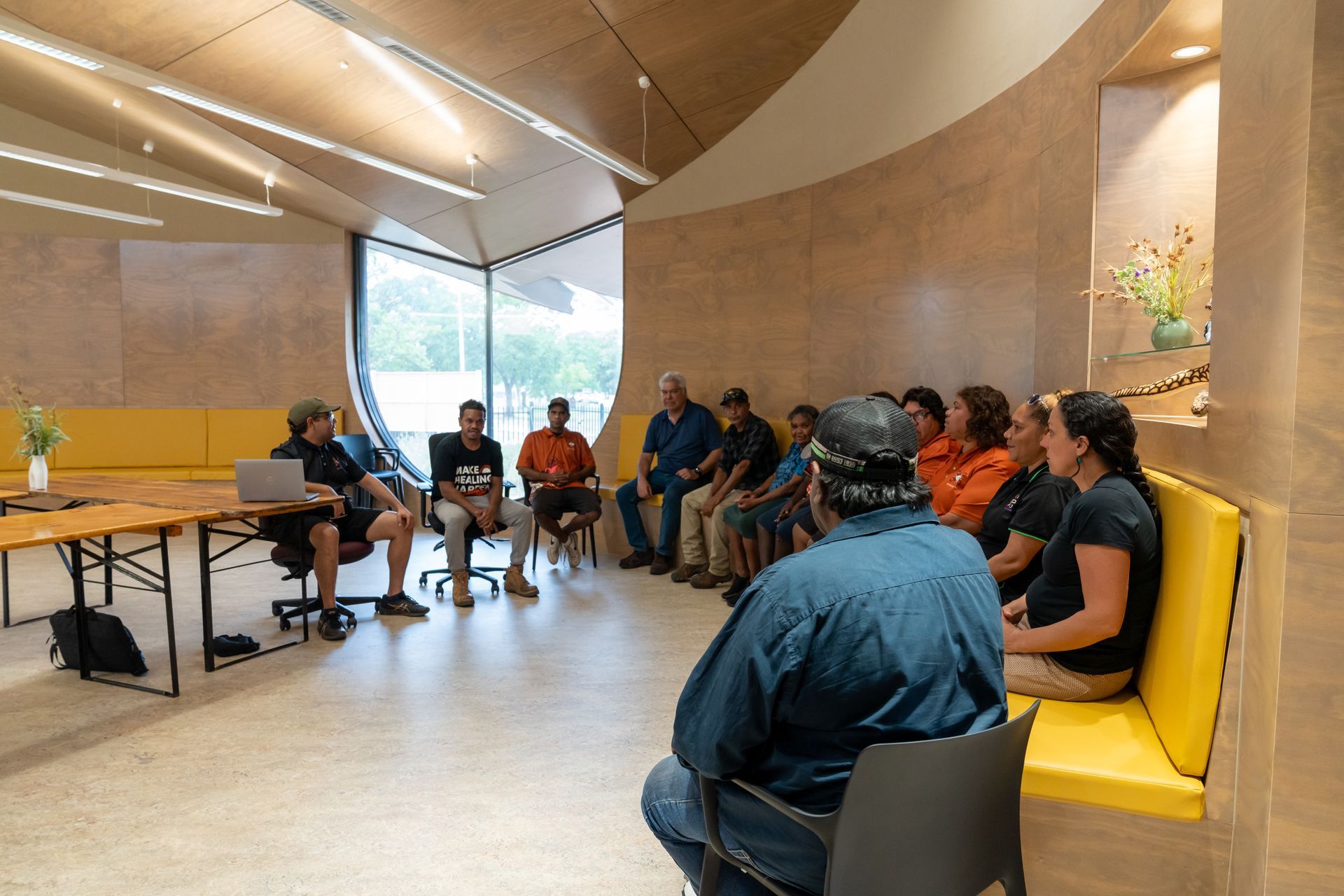 A group of people sit in a circle for a meeting inside a modern, wood-paneled room with yellow seating.