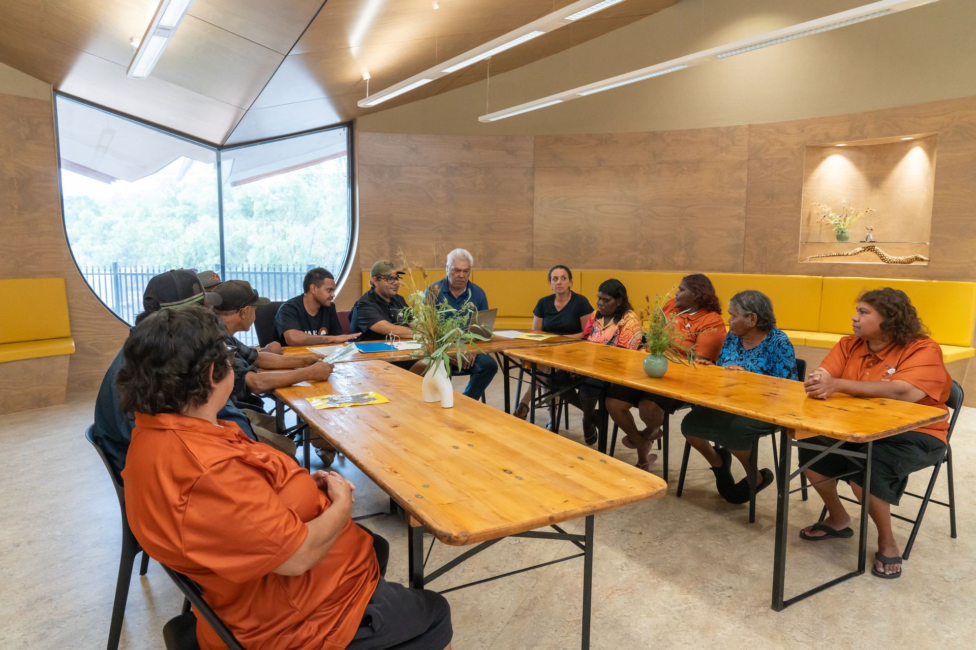 A group of people sit around long wooden tables in a bright, modern room with wooden walls for a meeting.