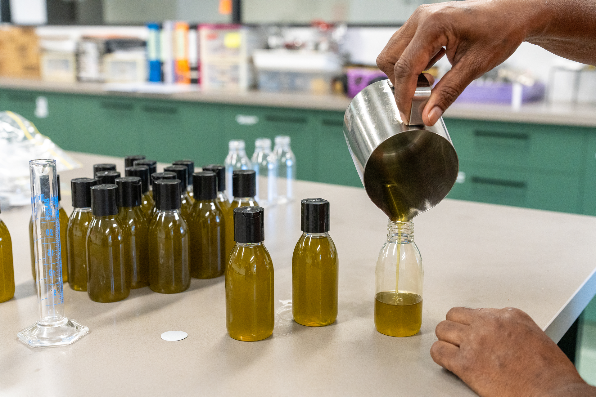 A person pours golden liquid from a metal pitcher into a small plastic bottle 