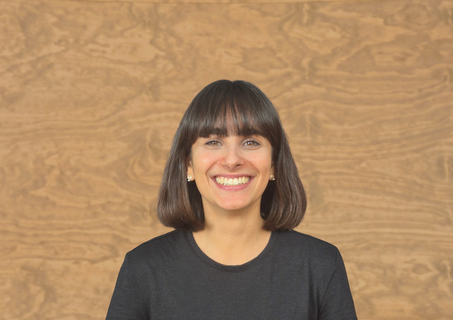 Smiling person with shoulder-length dark hair and bangs, wearing a black shirt, set against a wood-patterned background.