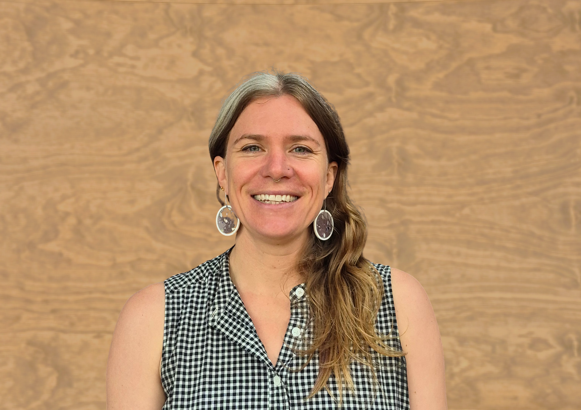 A smiling person wearing a black and white gingham sleeveless shirt and large, dangling earrings against a wood wall.