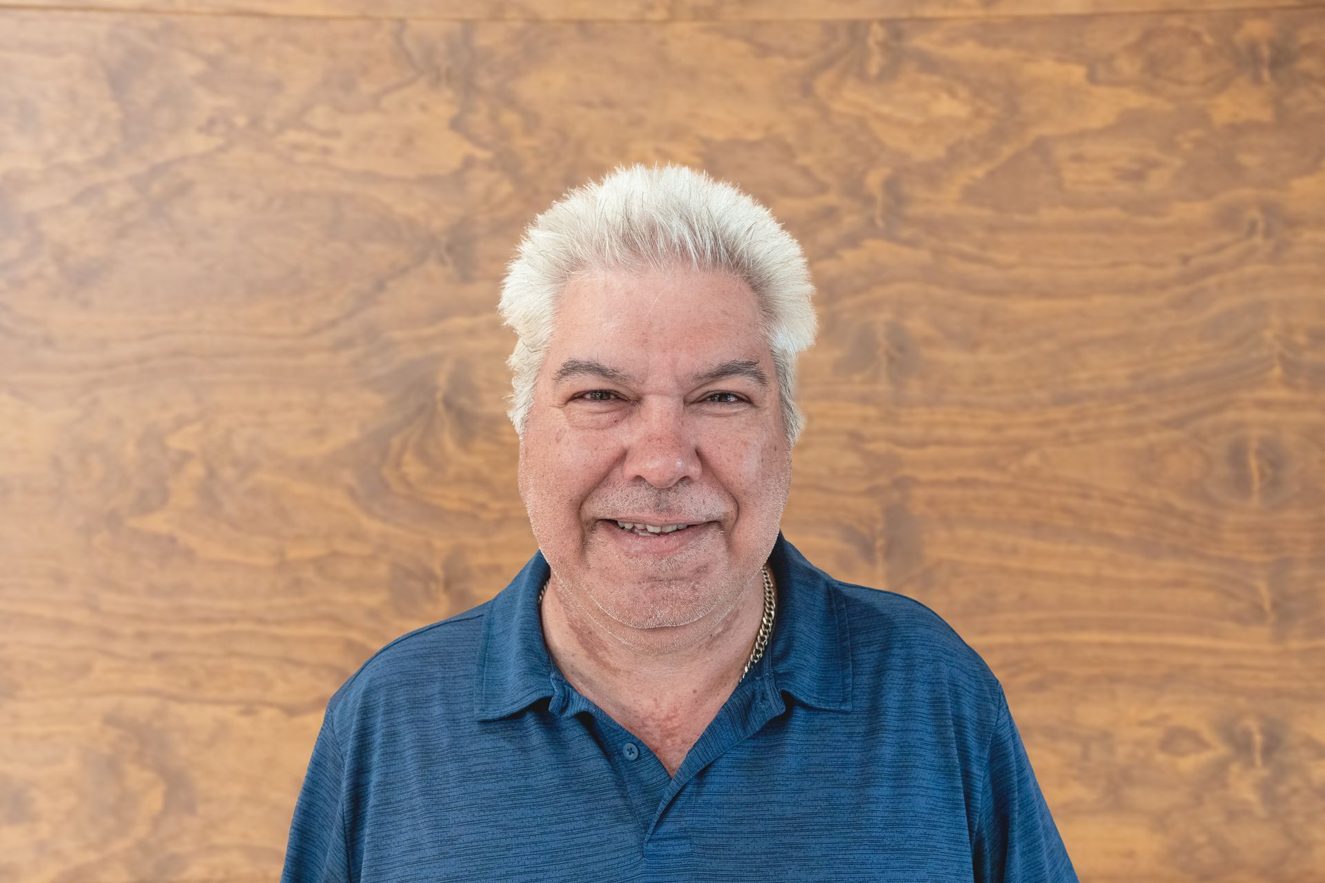 A person with white hair, wearing a blue polo shirt, smiling against a wood-paneled wall.