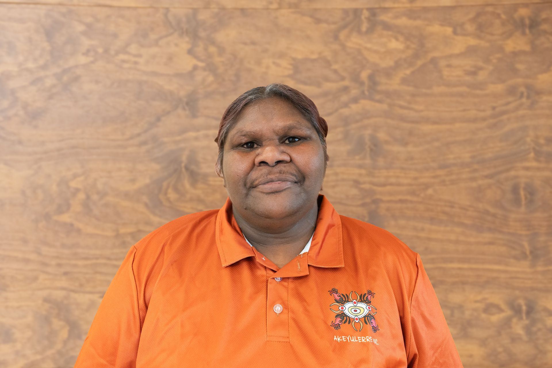 A person wearing an orange polo shirt with a small emblem, posing in front of a light brown wood-grain background.