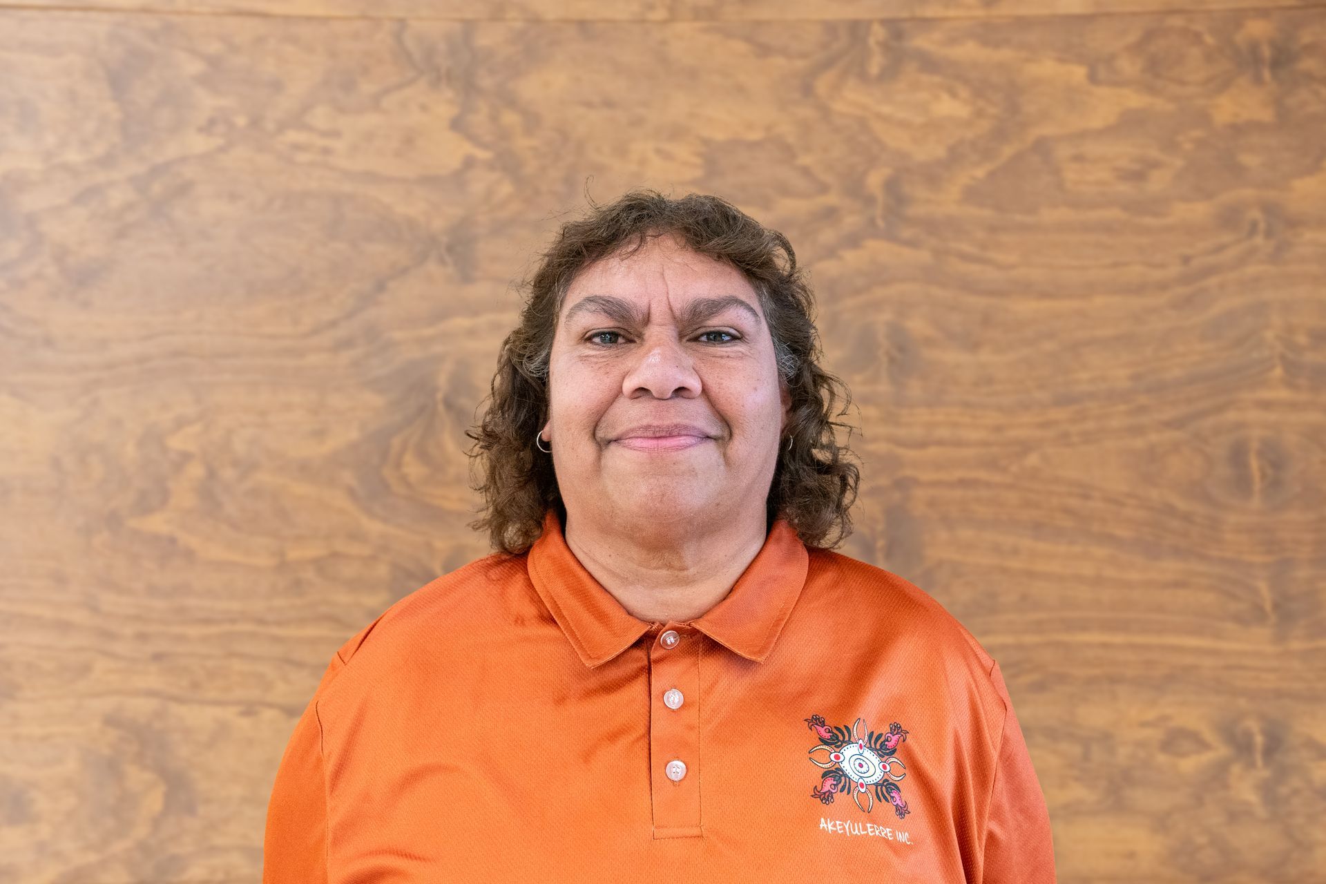 A person wearing a bright orange polo shirt with a logo, smiling against a light-brown wood-grain background.