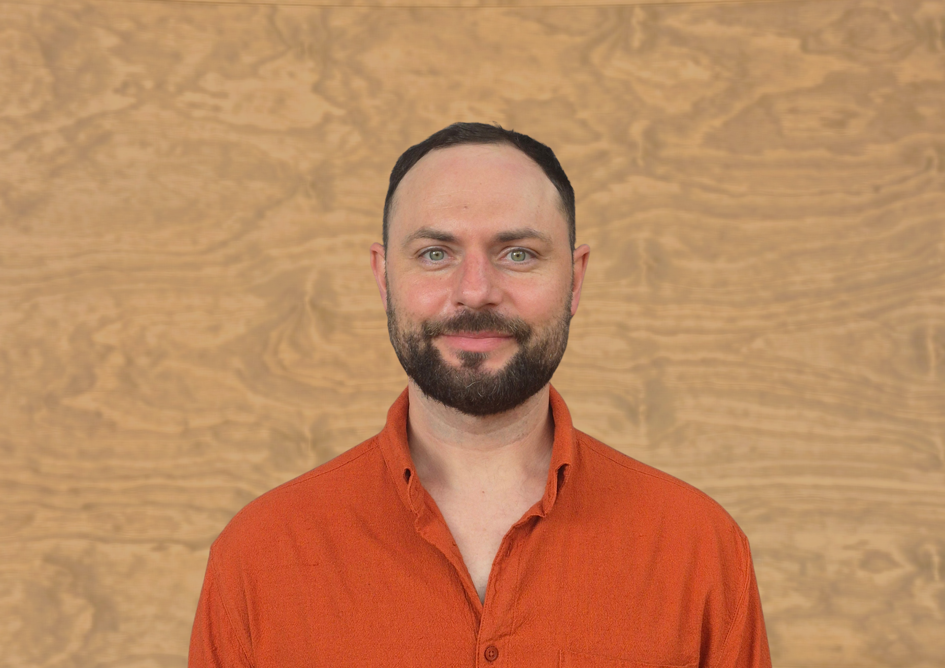 Man with a beard in an orange shirt smiling against a wood-paneled background