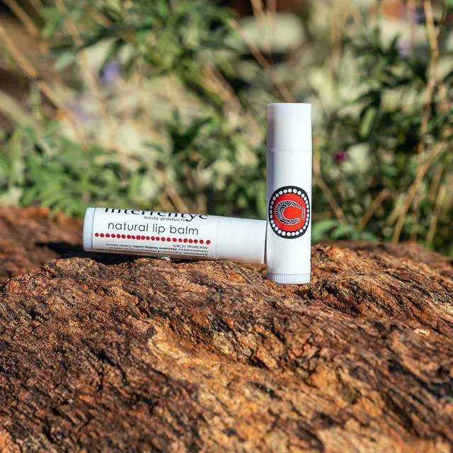 Two white lip balm tubes, branded with red circular logos and text, resting on a textured brown rock outdoors.