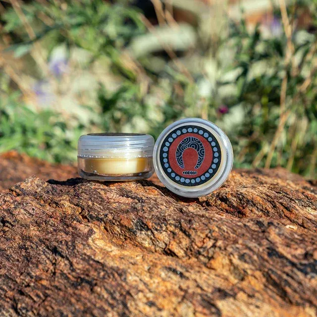 A small, clear jar of balm with a round decorative lid featuring a red circle and crescent icon on a textured rock.