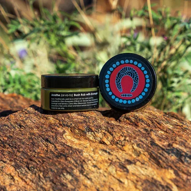 Two jars of dark green balm with ornate, colorful circular lids resting on a rugged, textured rock in an outdoor setting.