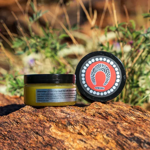 A small jar of skincare product with an Aboriginal-inspired circular logo, resting on a textured rock in a desert setting.