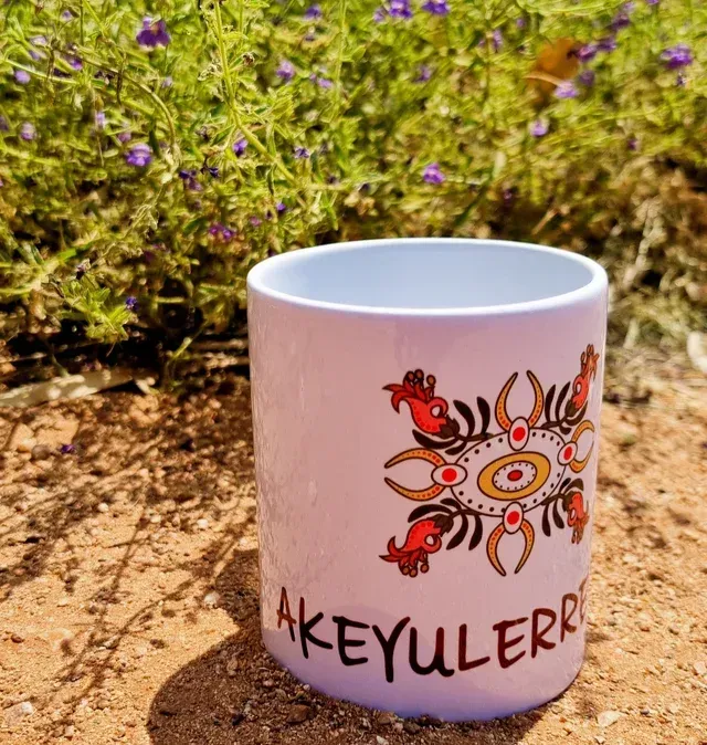 A white mug with indigenous-style orange and red artwork, sitting on reddish-brown earth near small purple wildflowers.