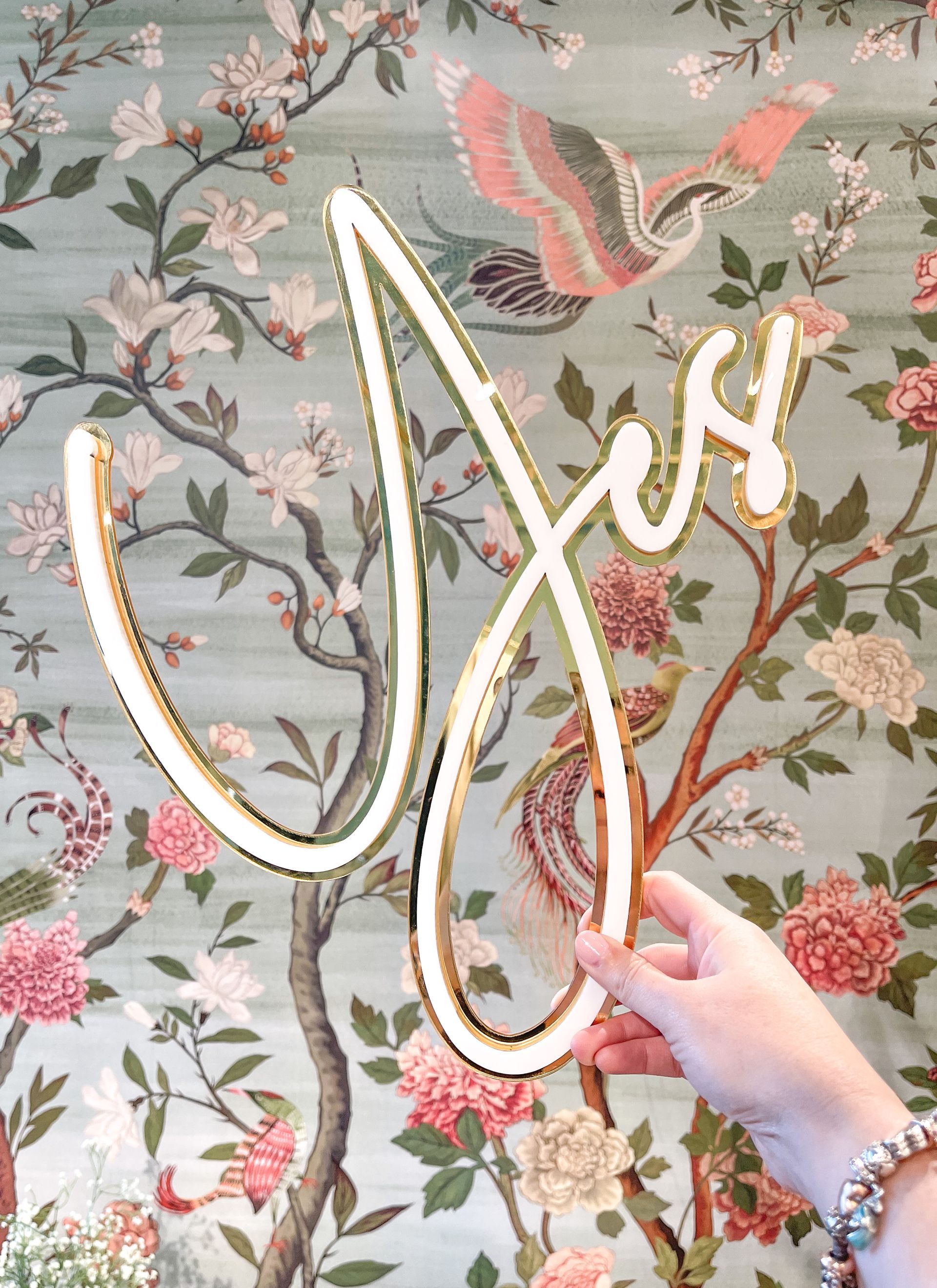 A person is holding a neon sign in front of a floral wallpaper.