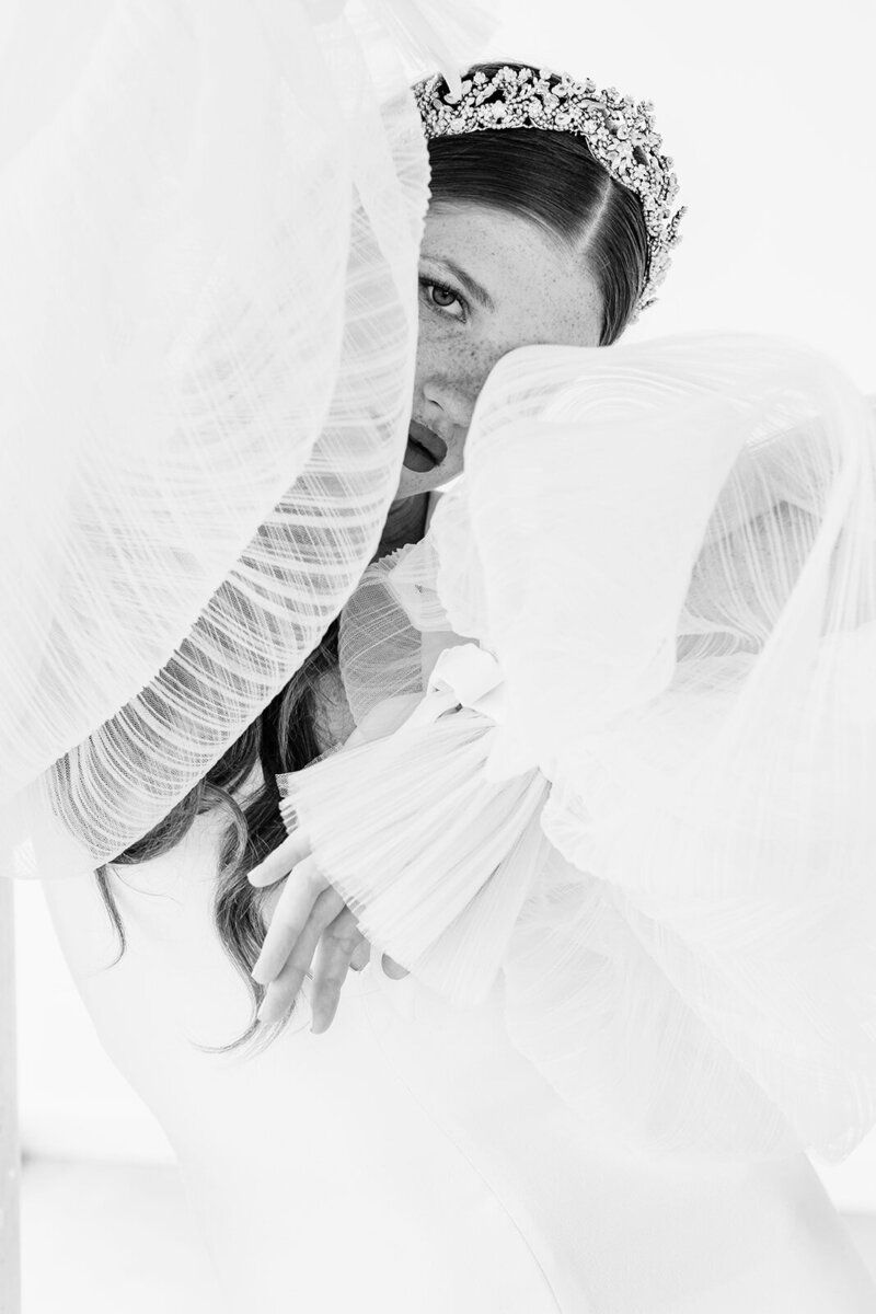 A black and white photo of a bride in a wedding dress covering her face with a veil.