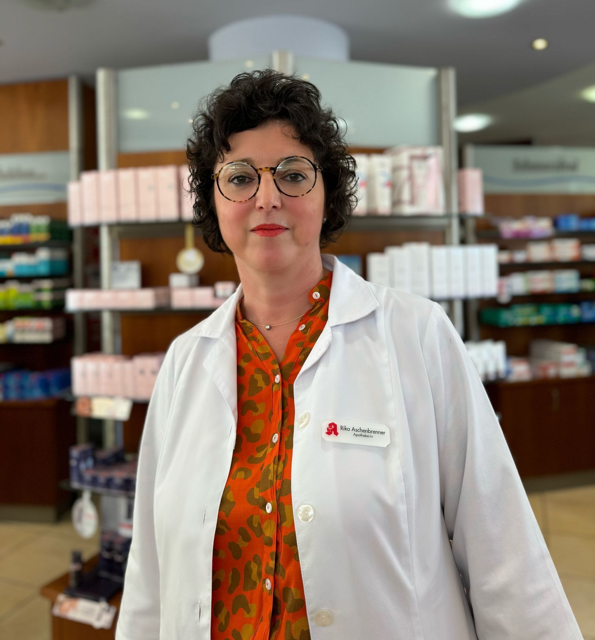 Eine Frau mit Brille und weißem Kittel steht in einer Apotheke.