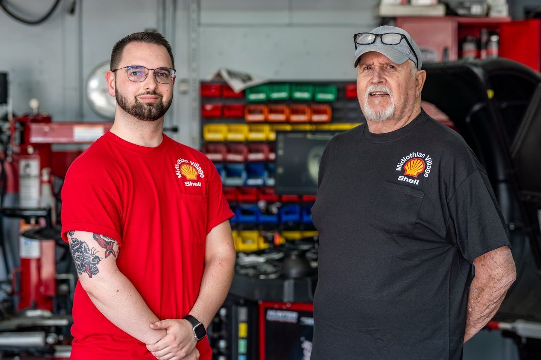 Two men in auto shop, wearing red/black shirts. One with glasses and beard, other with cap. Shelves with parts visible.