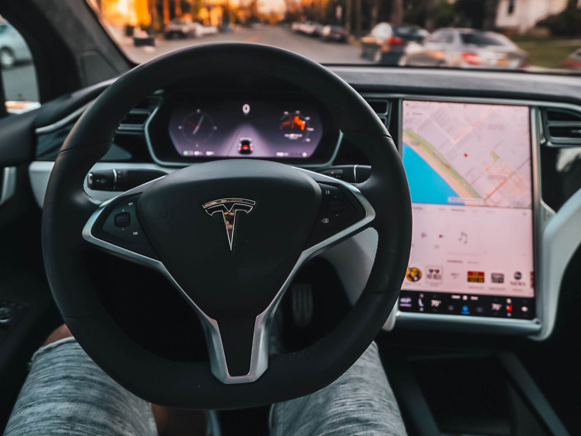 Dashboard view of a Tesla car interior, with a touchscreen display and driver's hands on the steering wheel.