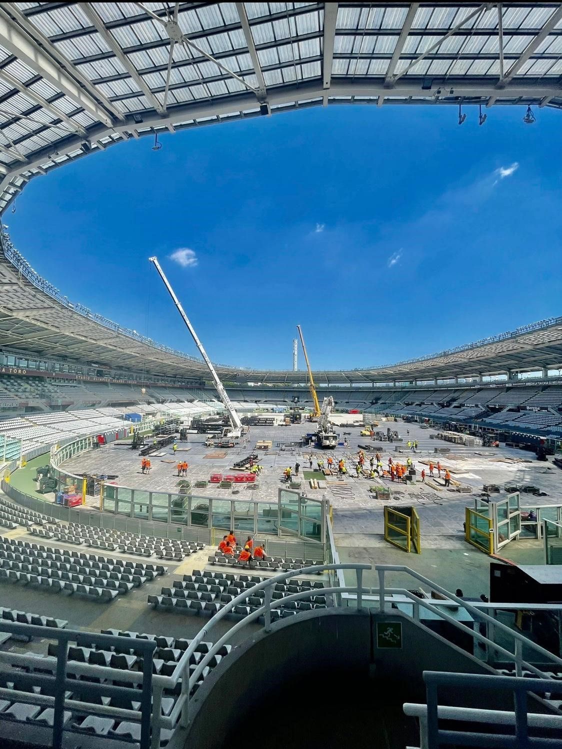 Cantiere all'interno di uno stadio. Operai e gru sul campo, sotto un cielo azzurro brillante.