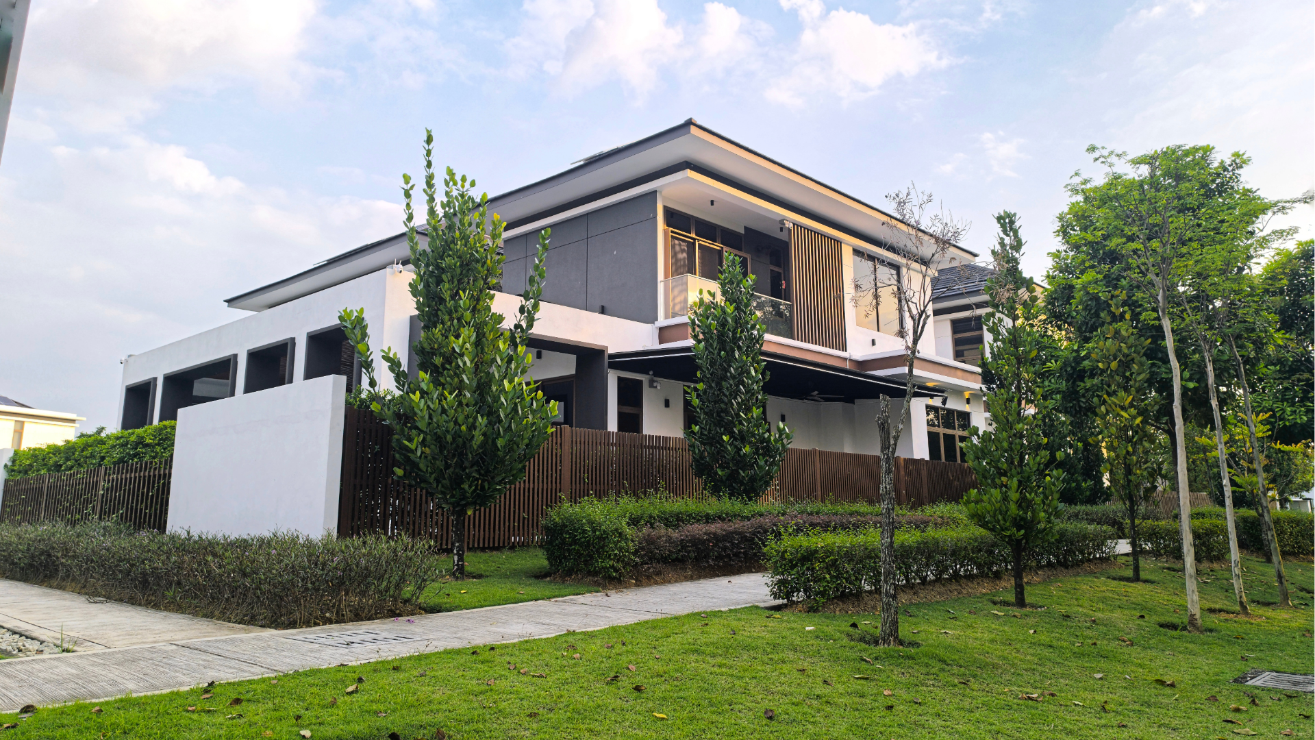 Modern two-story house with a balcony, surrounded by green lawn and trees under a cloudy sky