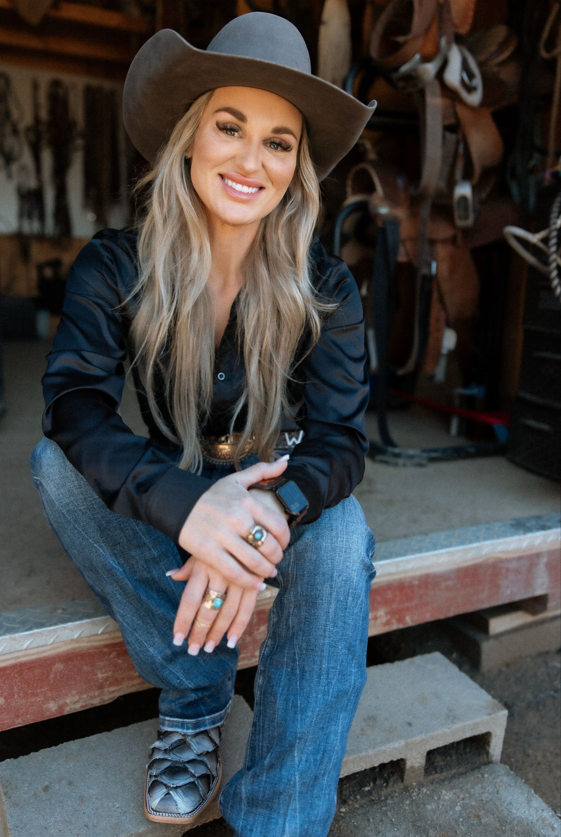 A woman wearing a cowboy hat and jeans is sitting on the steps of a stable.