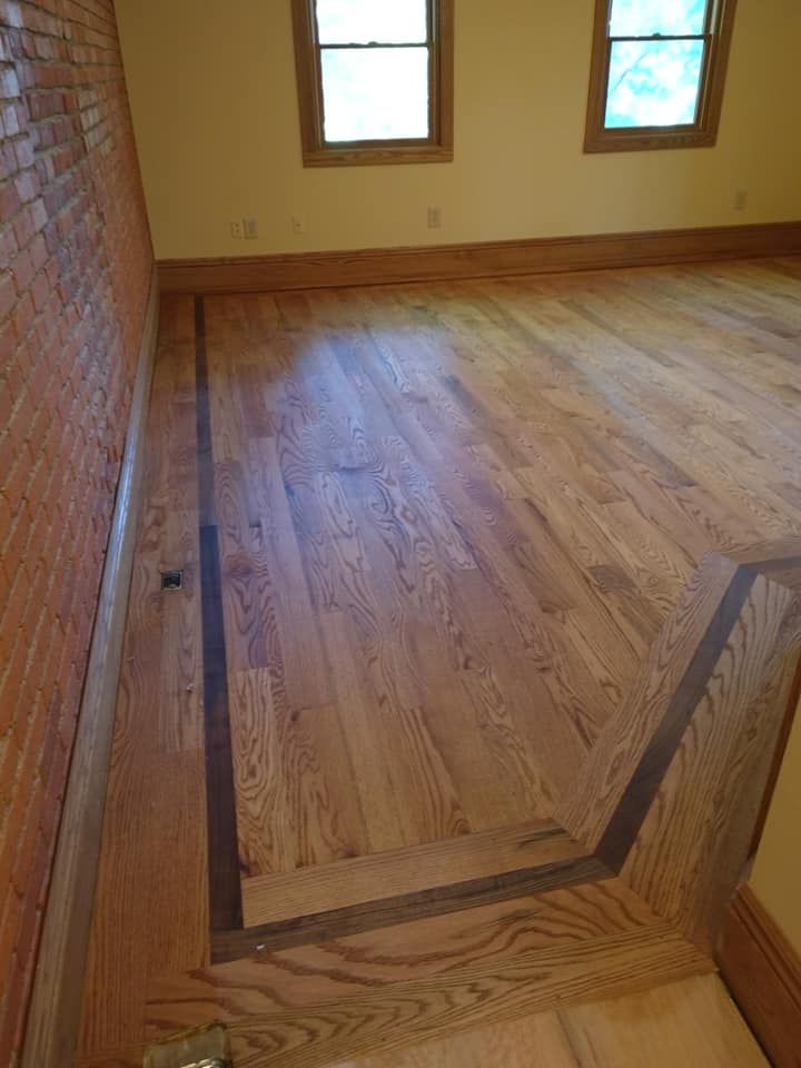Wooden floor with dark border, next to a brick wall. Two windows in the background.