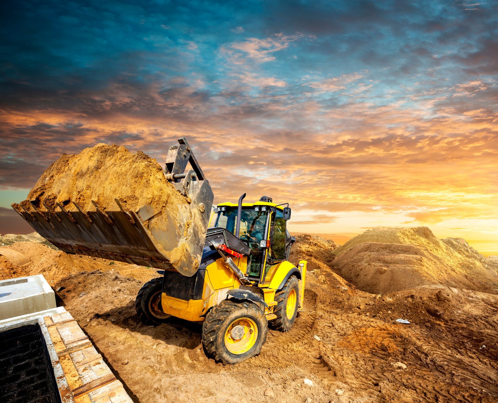 Una pala meccanica gialla scarica terra in un cantiere edile con cumuli di terra sotto un cielo spettacolare.
