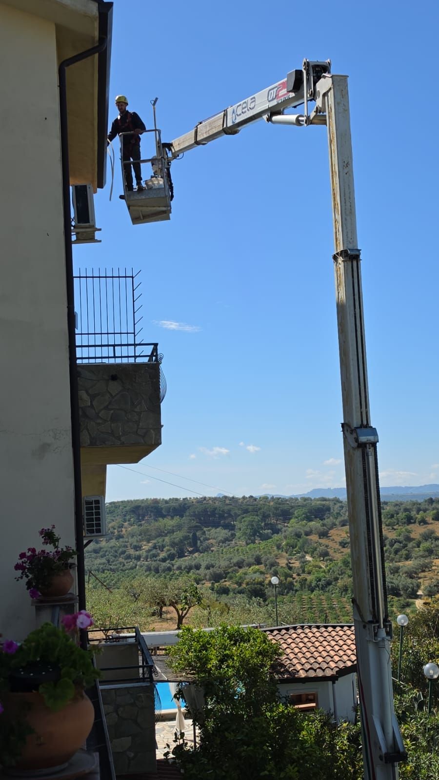 Un operaio a bordo di una piattaforma aerea ispeziona la facciata di un edificio sullo sfondo di uno scenario collinare suggestivo.