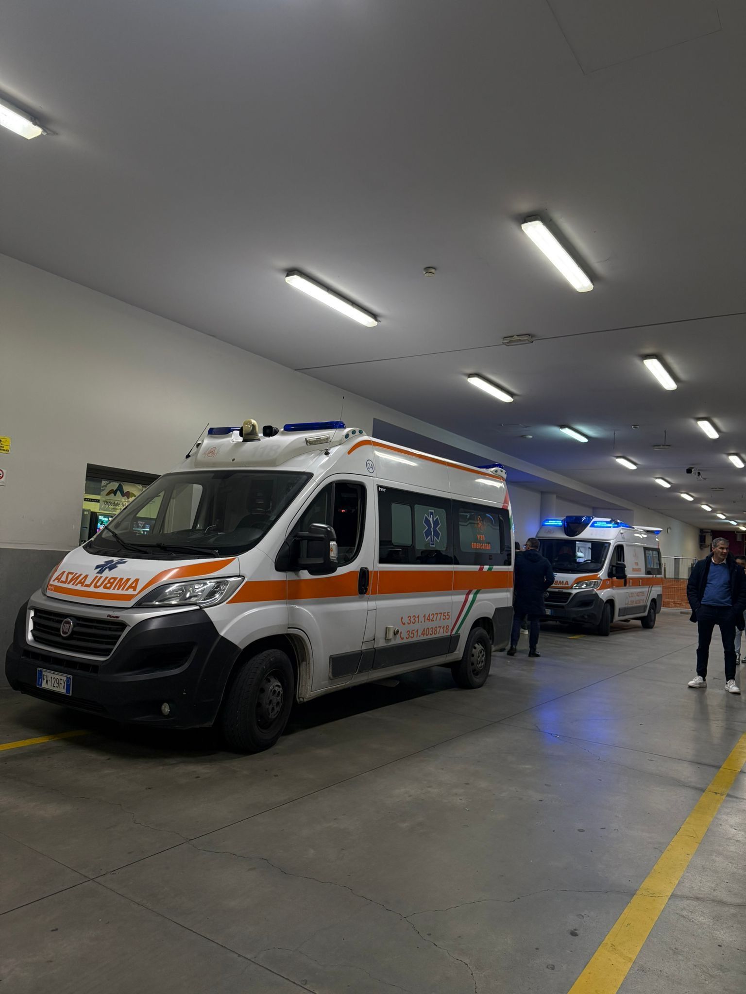 Ambulanza parcheggiata in un garage coperto con persone nelle vicinanze e luci accese.