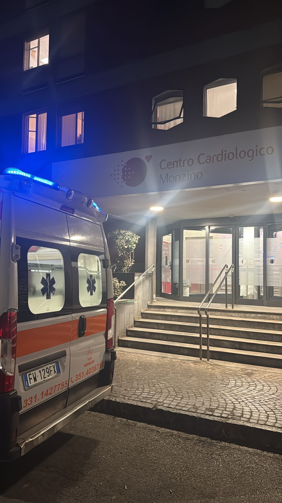 Un'ambulanza parcheggiata di notte davanti all'ingresso illuminato dell'ospedale, con una scala che conduce a porte a vetri.