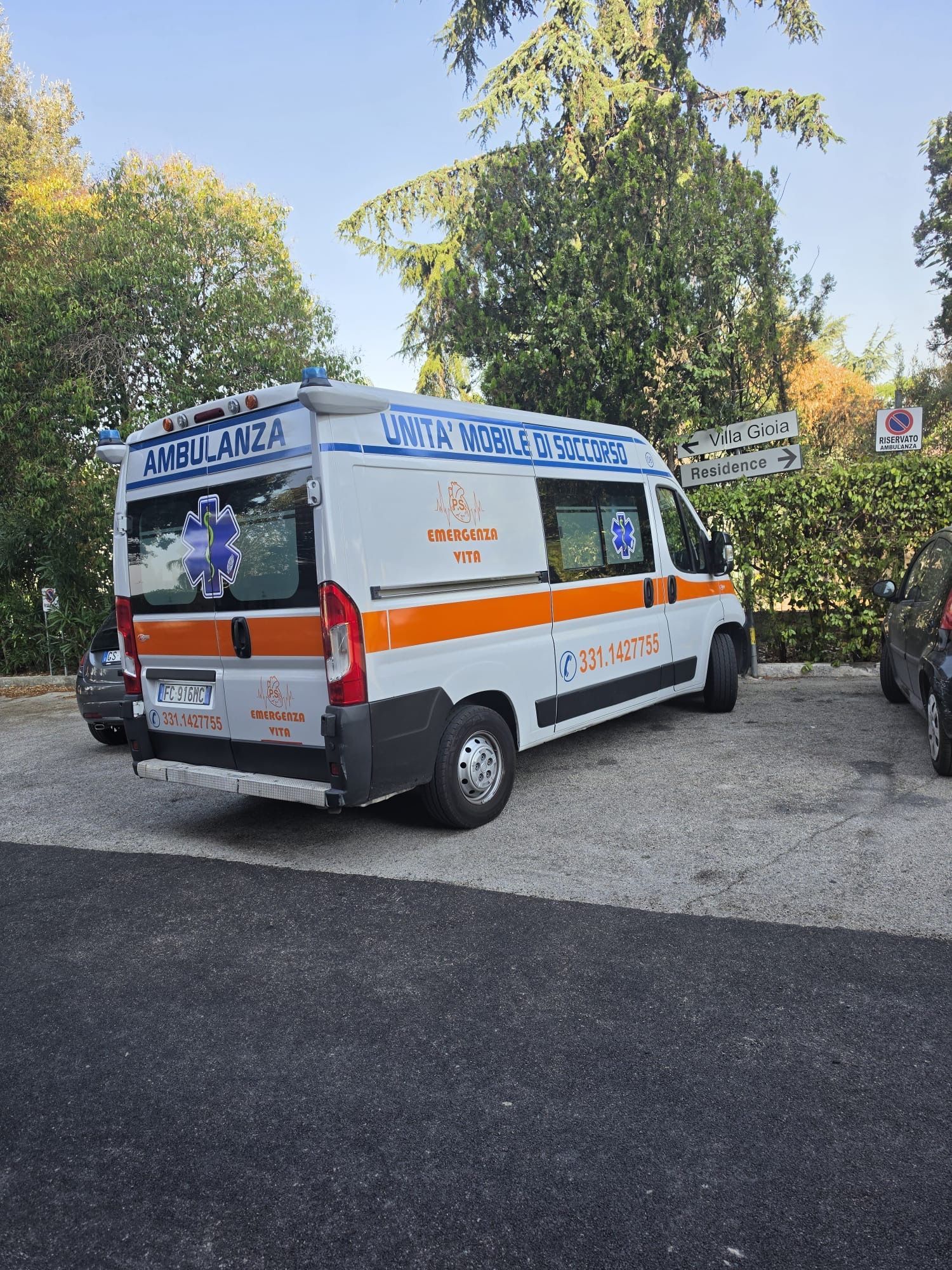 Un furgone ambulanza bianco con una striscia arancione parcheggiato all'aperto sotto gli alberi, con luce blu e segnaletica sul tetto.