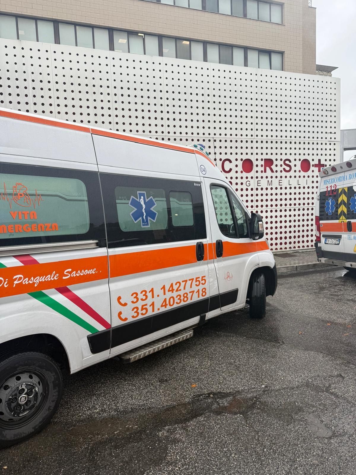 Ambulanza bianca con striscia arancione parcheggiata fuori da un edificio ospedaliero.