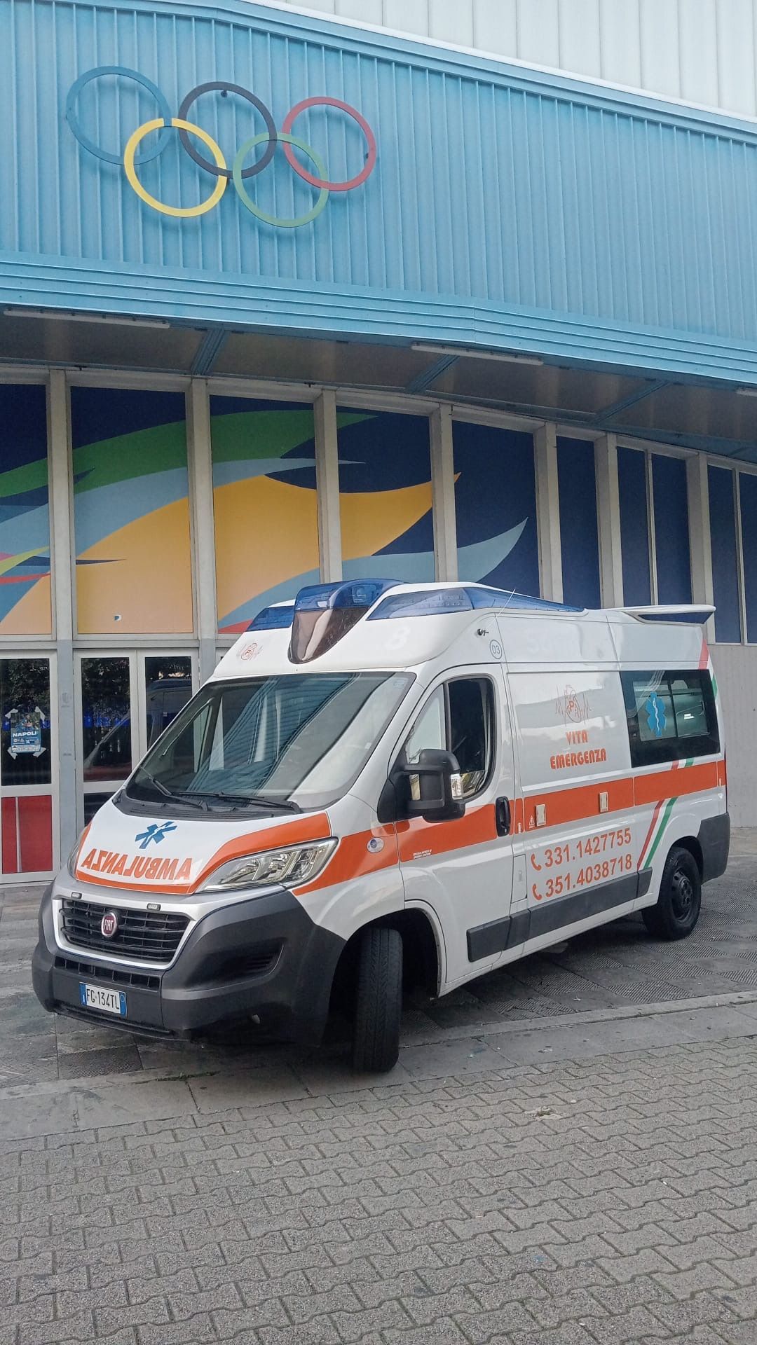 Un'ambulanza bianca e arancione con il logo degli anelli olimpici è parcheggiata fuori da un impianto sportivo.