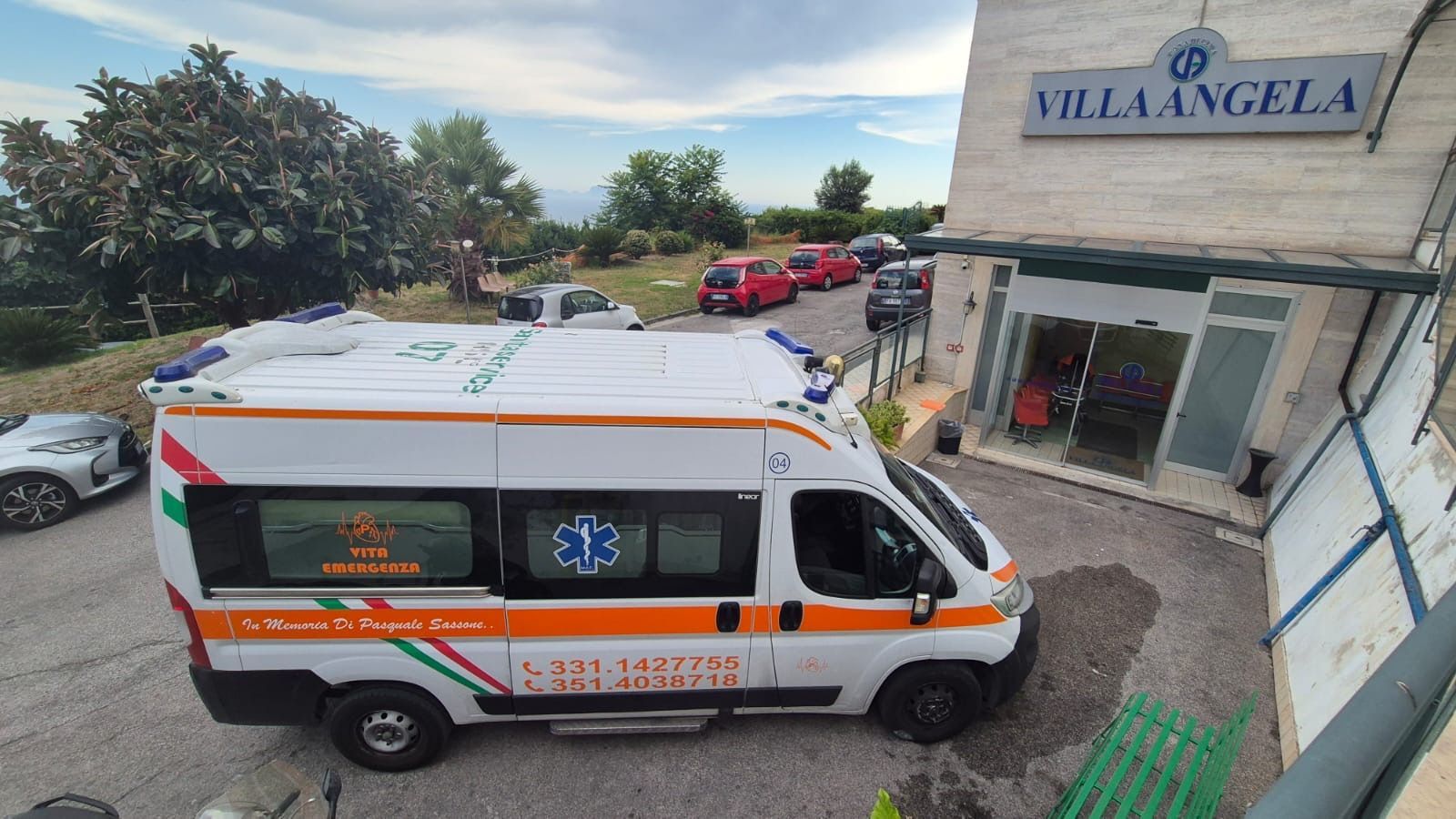 Un'ambulanza è parcheggiata all'esterno dell'ingresso di Villa Angela, con auto nel parcheggio e l'insegna dell'edificio ben visibile.