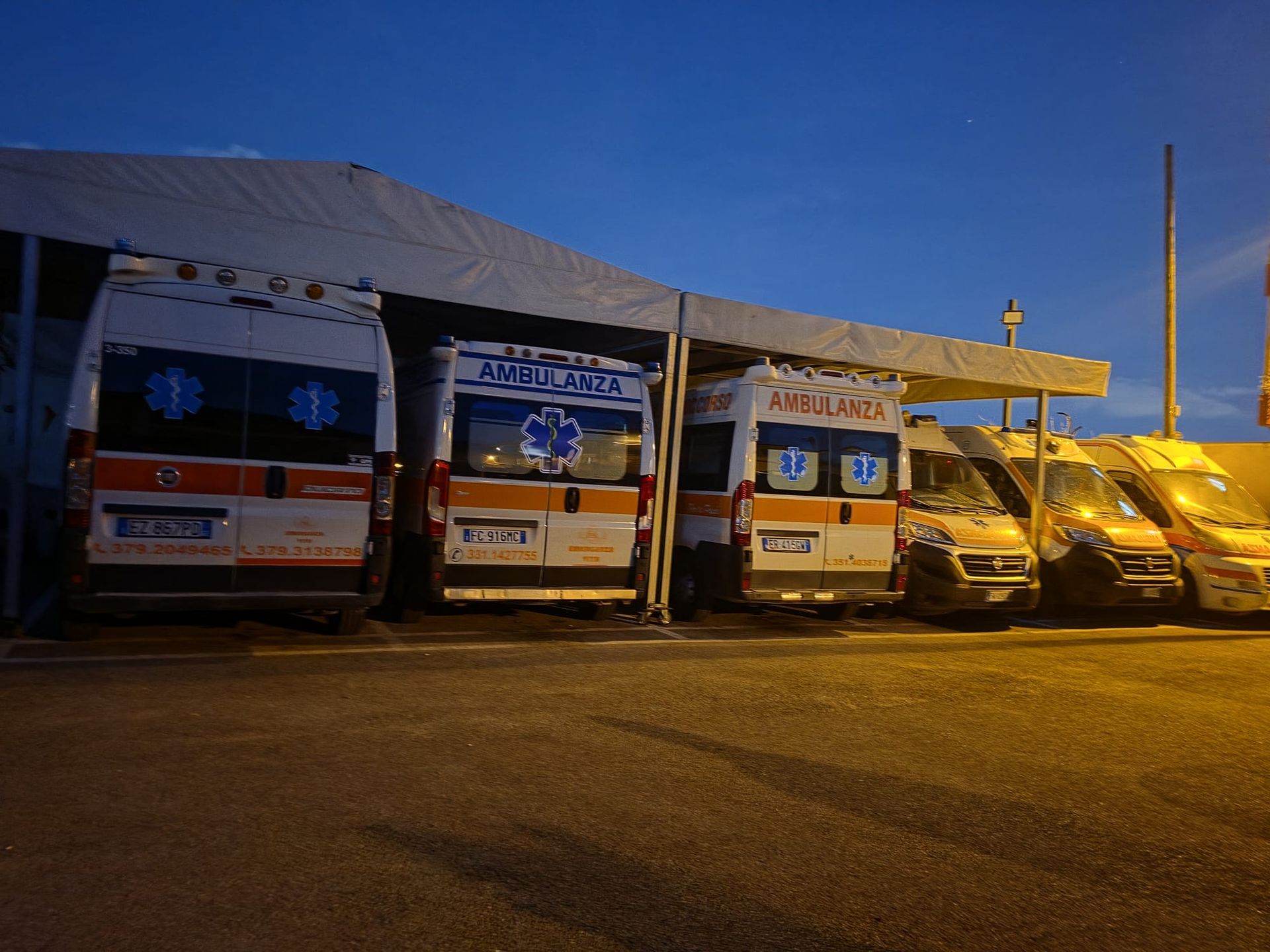 Ambulanze e veicoli di emergenza parcheggiati e allineati accanto a una tenda illuminata al crepuscolo.