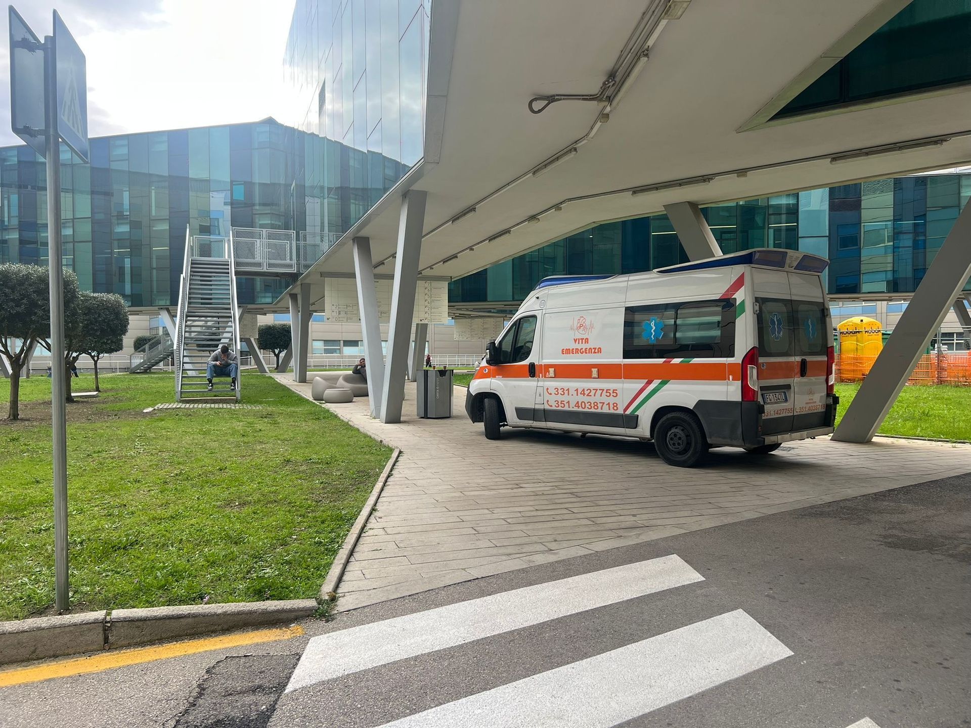 Ambulanza parcheggiata sotto la sporgenza di un edificio moderno, accanto a un attraversamento pedonale e a uno spartitraffico erboso.