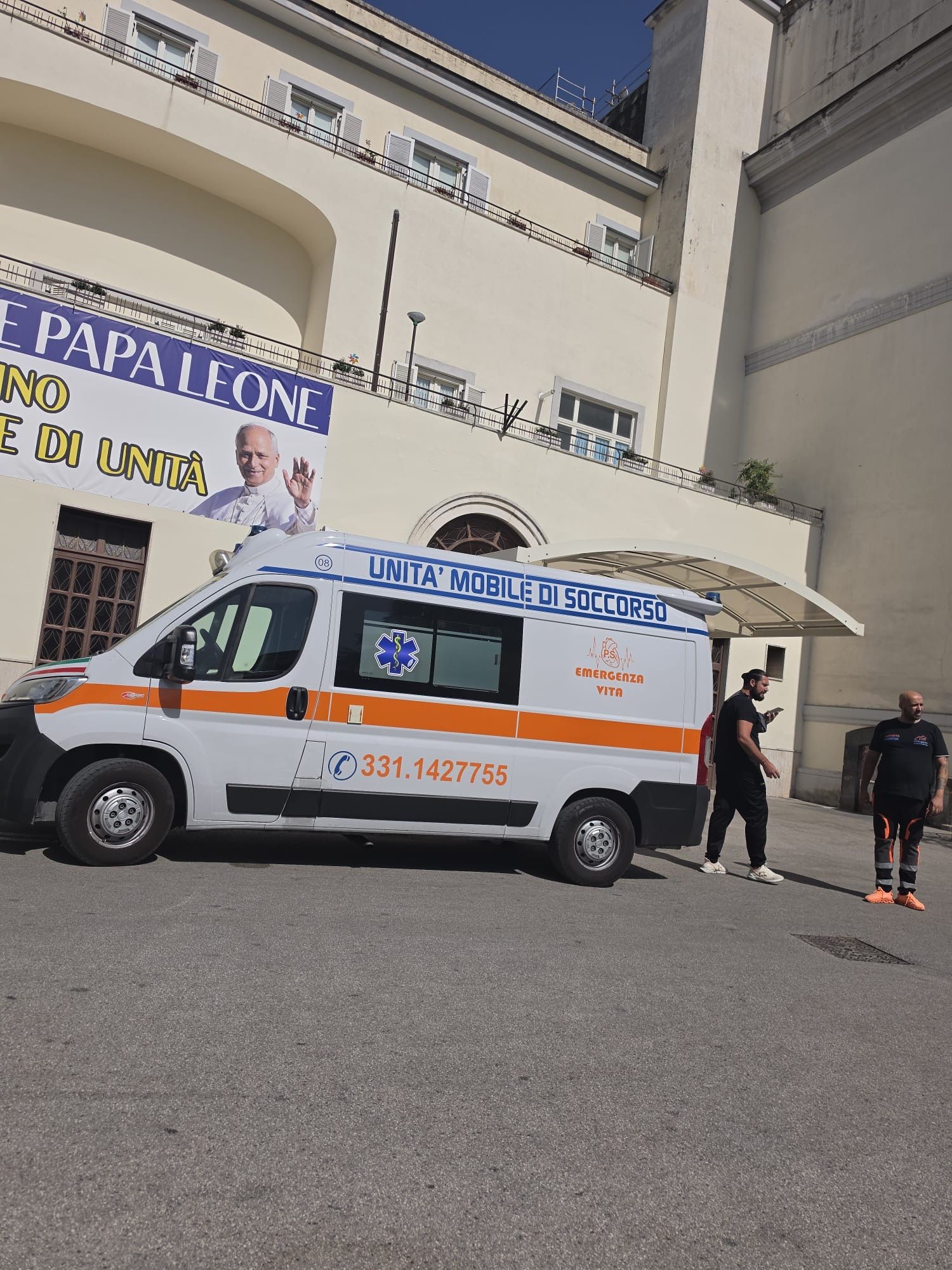 Un'ambulanza bianca e arancione è parcheggiata fuori da un edificio, con due persone in piedi nelle vicinanze.