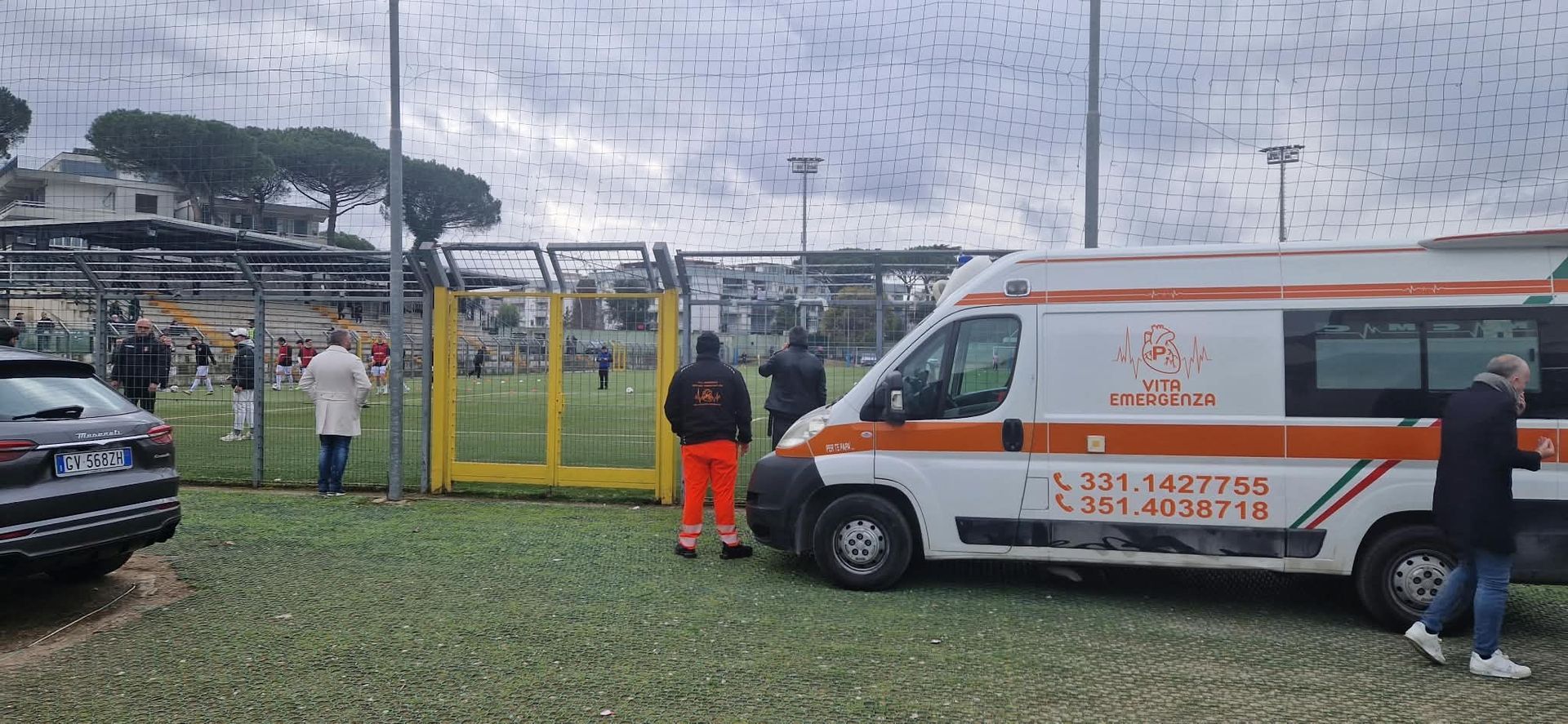 Un'ambulanza è parcheggiata vicino a un campo sportivo recintato, con persone in piedi nelle vicinanze, in una giornata nuvolosa.