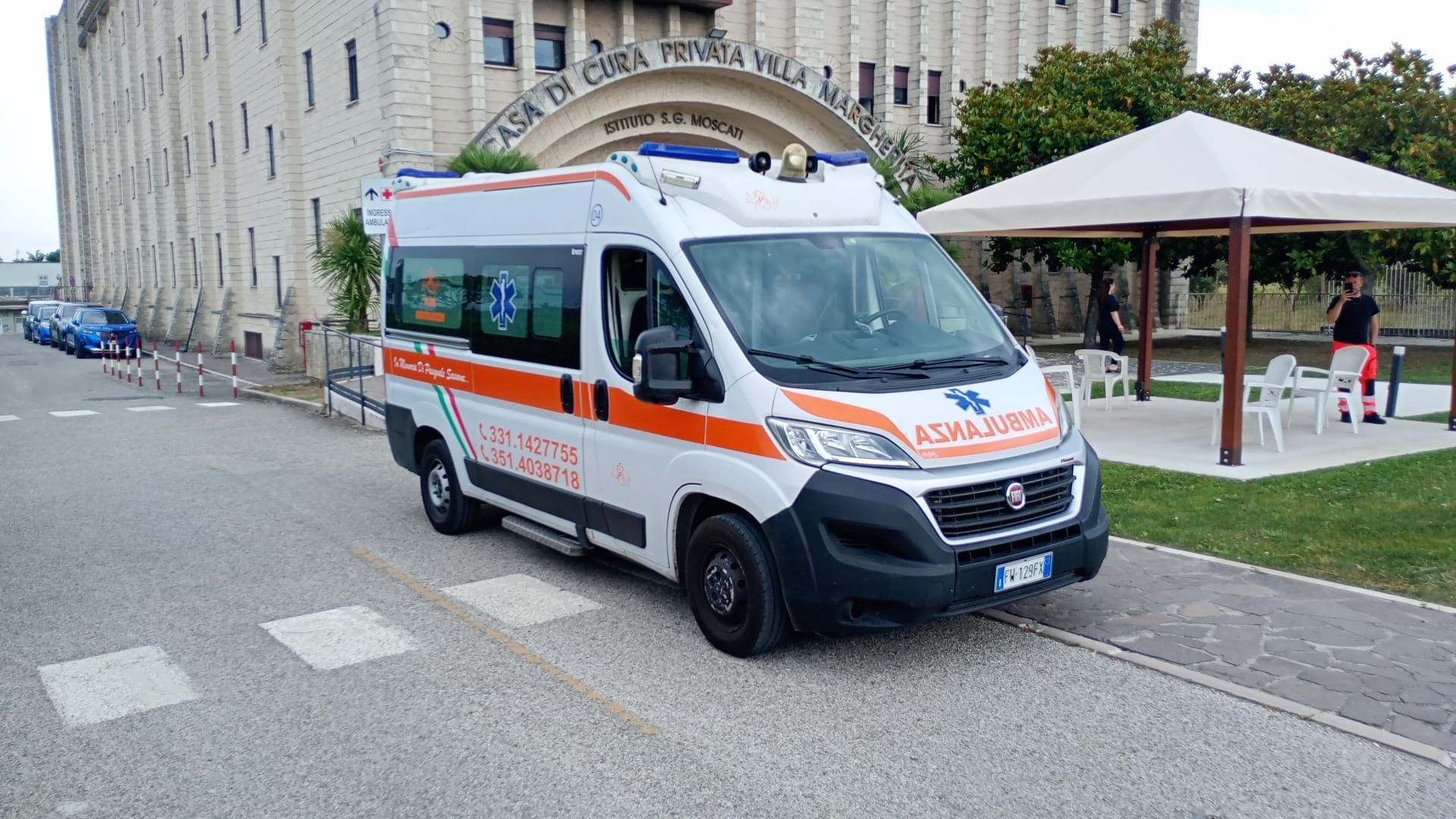 Un'ambulanza è parcheggiata davanti all'ingresso di un edificio, con una persona nelle vicinanze riparata sotto una tettoia bianca.