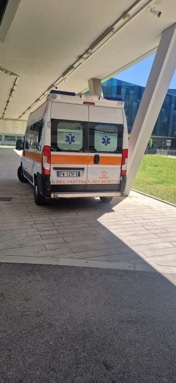Ambulanza parcheggiata sotto un vialetto coperto, portelloni posteriori visibili con segnaletica di emergenza rossa e blu.