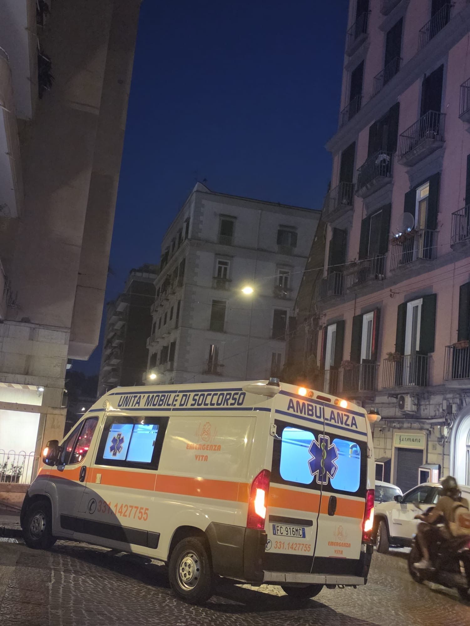 Un'ambulanza con le luci lampeggianti in una stretta via cittadina di notte, circondata da alti edifici.