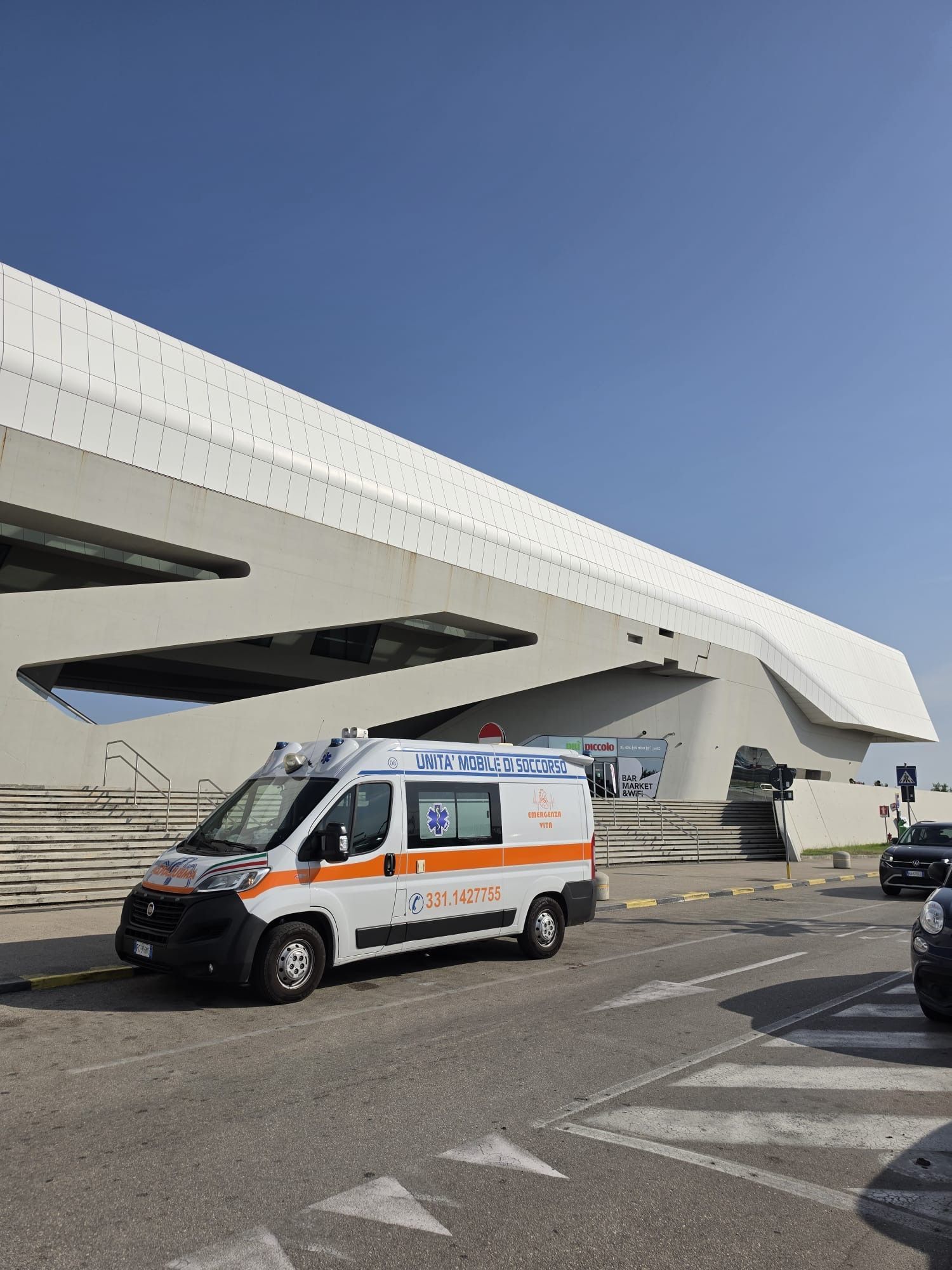Ambulanza parcheggiata accanto a un grande cavalcavia in cemento su una strada soleggiata.