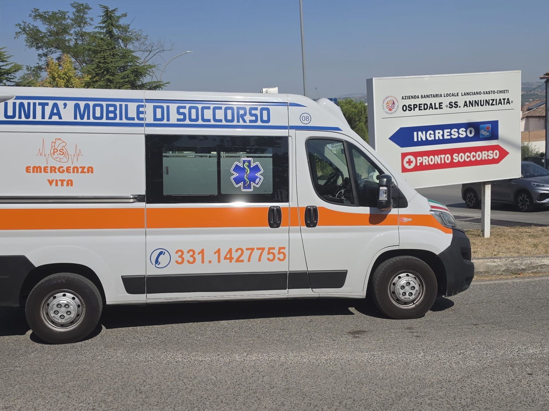 Ambulance van with “Unità Mobile di Soccorso” parked beside a roadside sign for emergency medical services.