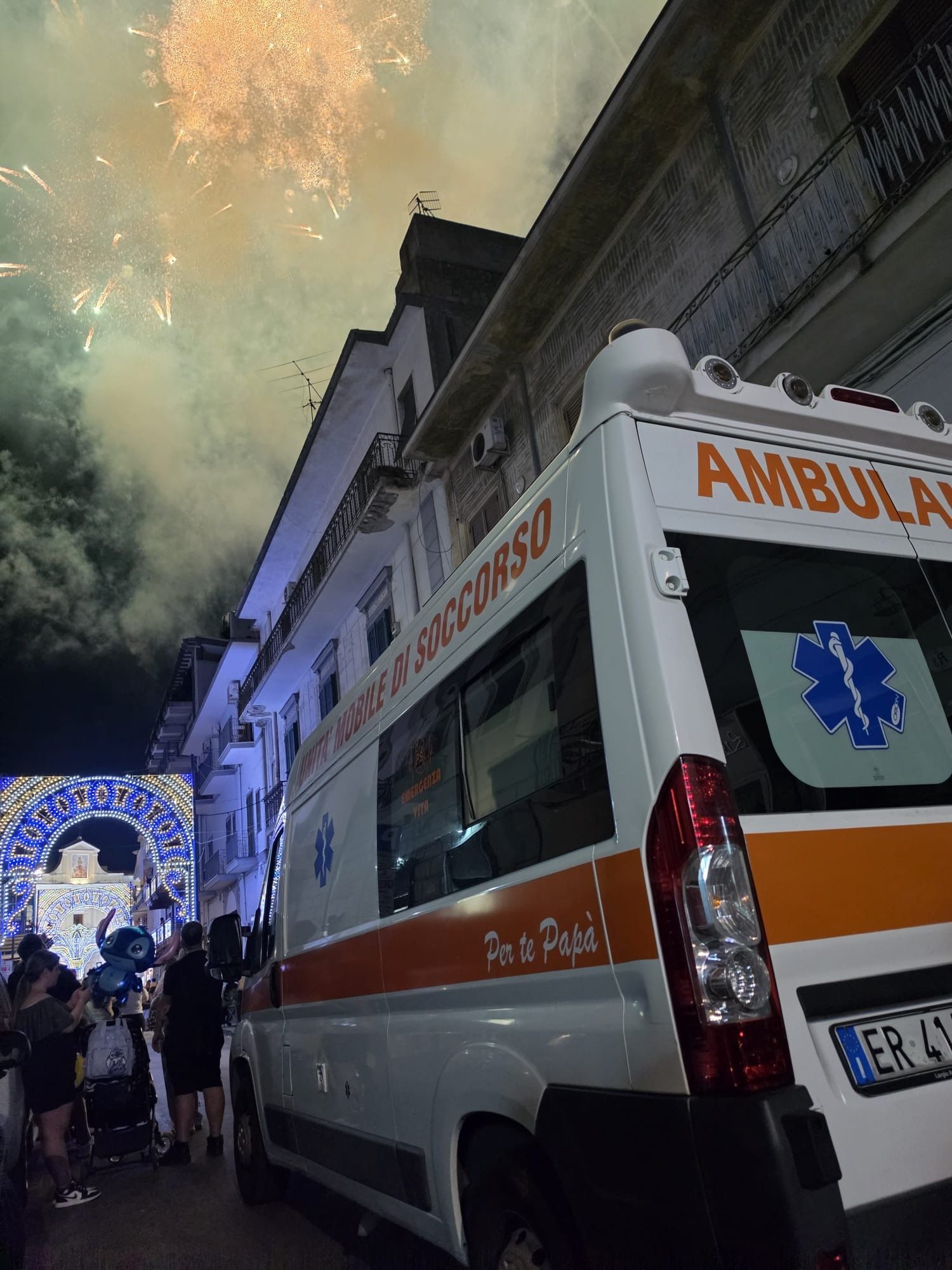 Un'ambulanza parcheggiata di notte in una strada stretta, sotto i fuochi d'artificio, con un arco di festa illuminato sullo sfondo.