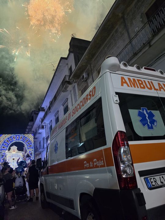 Un'ambulanza parcheggiata in una strada stretta accanto a una folla illuminata, sotto un cielo notturno fumoso.